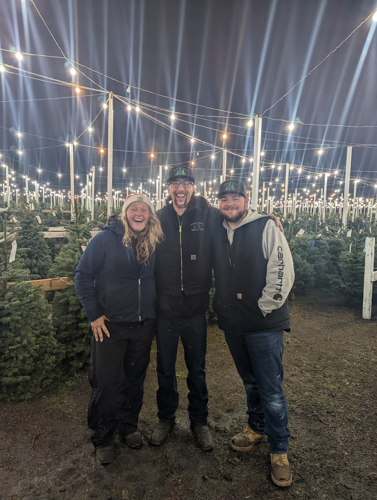 Owner Morgan Gutzman with elves Jacob and Mitchell, laughing together on the lot at night under strings of lights