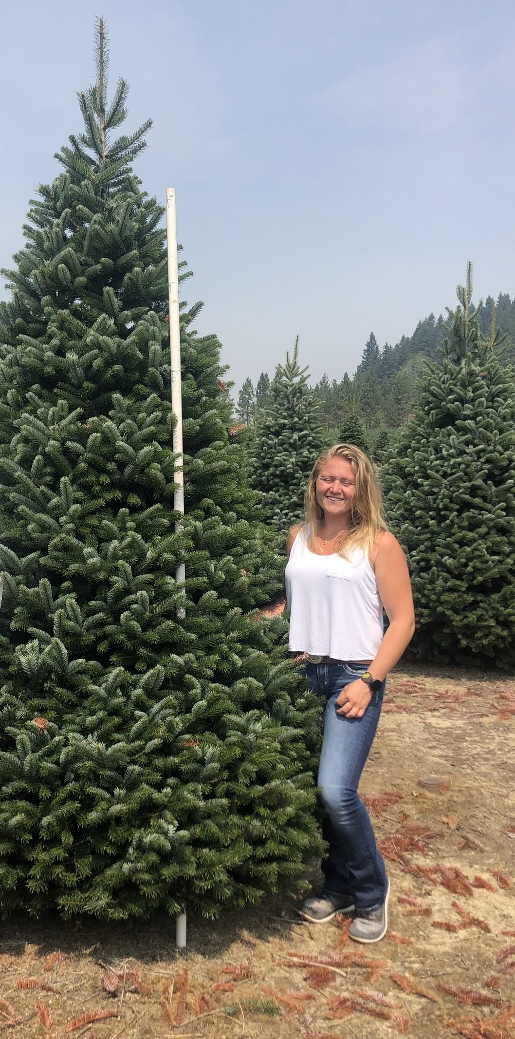 Morgan Gutzman, in a white tank top and jeans, smiling beside a tall fir with a measuring pole at a tree farm