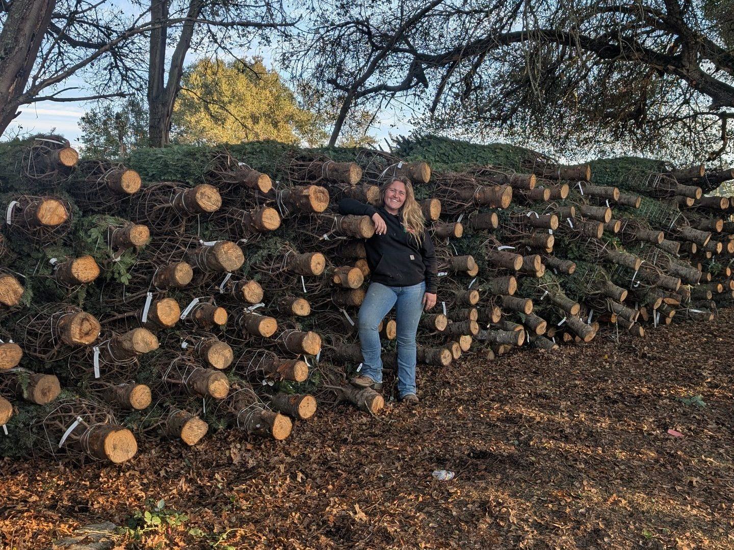 Morgan Gutzman standing in front of a huge pile of freshly cut Christmas trees